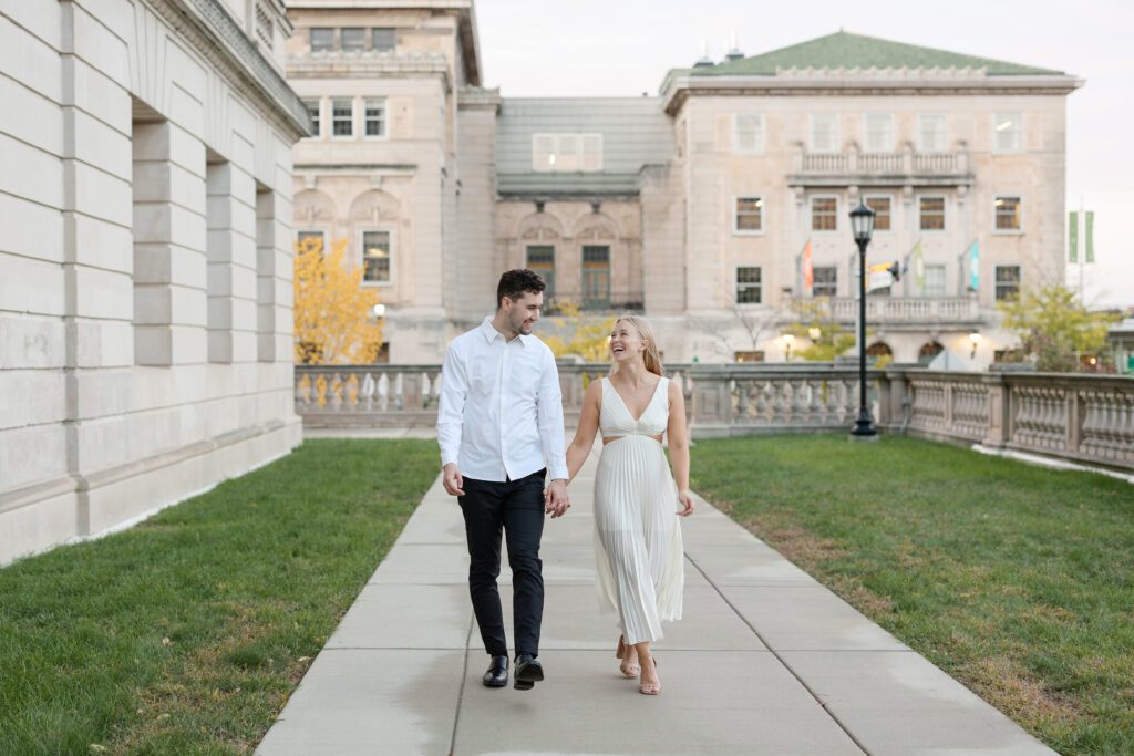 Engaged couple holding hands and walking together with Memorial Union views in the background.