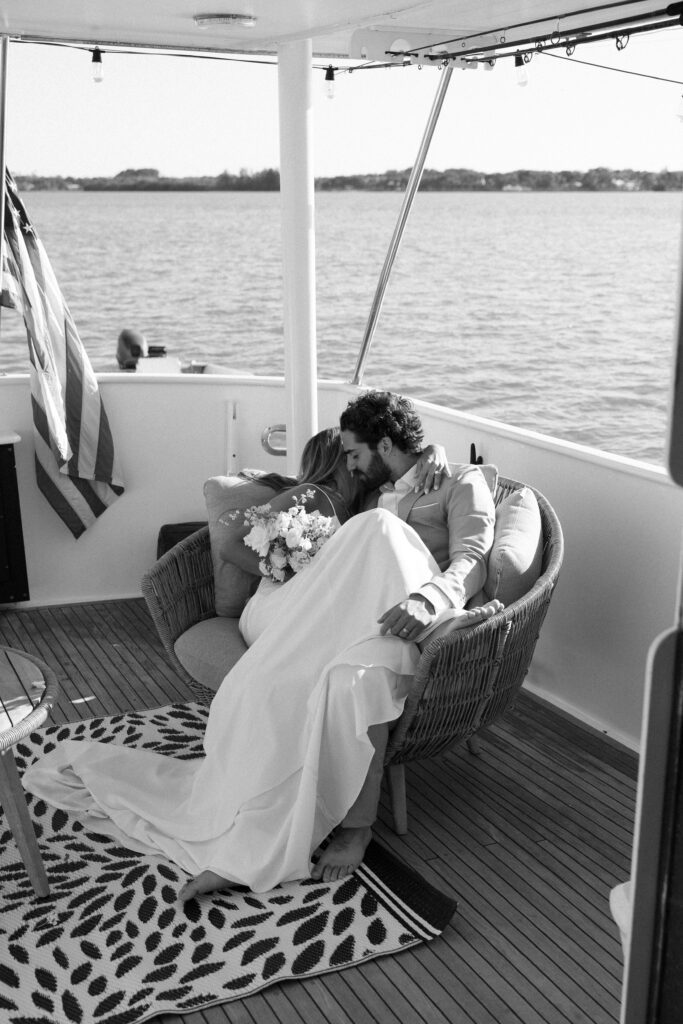 The bride and groom snuggle close on a chair, enjoying a cozy moment together aboard their yacht during the West Palm Beach Boat Elopement.