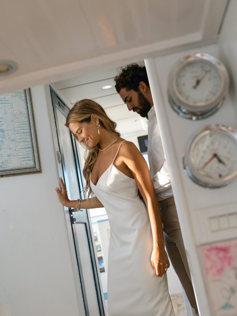 The groom carefully zips up the bride's dress in the yacht cabin, capturing a tender moment before their West Palm Beach Boat Elopement.