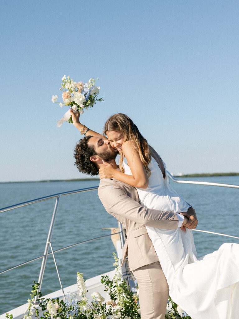 The bride joyfully throws her bouquet into the air while the groom lifts her up on the stern of the boat, celebrating their West Palm Beach Boat Elopement.