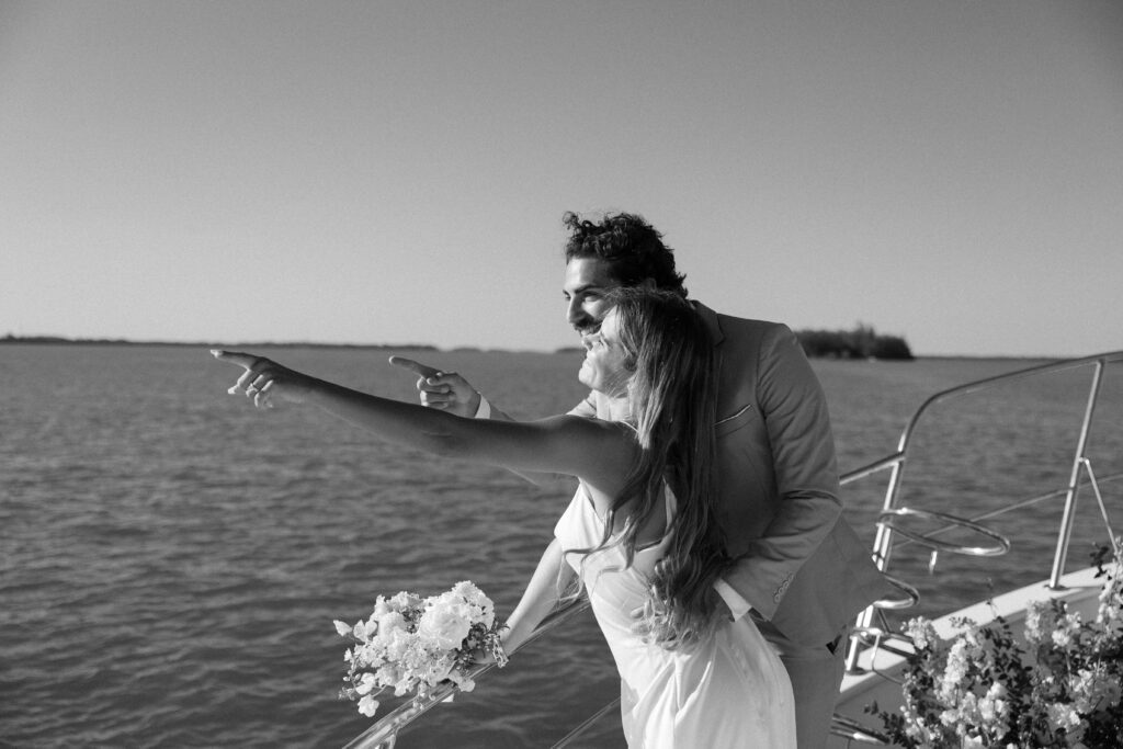 The couple smiles and points off into the distance from the deck of their yacht, capturing the joy of their West Palm Beach Boat Elopement.